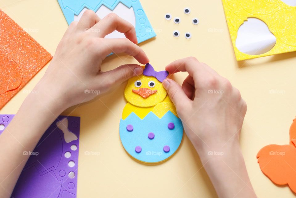 The hands of a caucasian teenage girl glue a lilac felt bow on a yellow chicken, sitting at the children's table, close-up side view. Concept diy, Easter preparation.