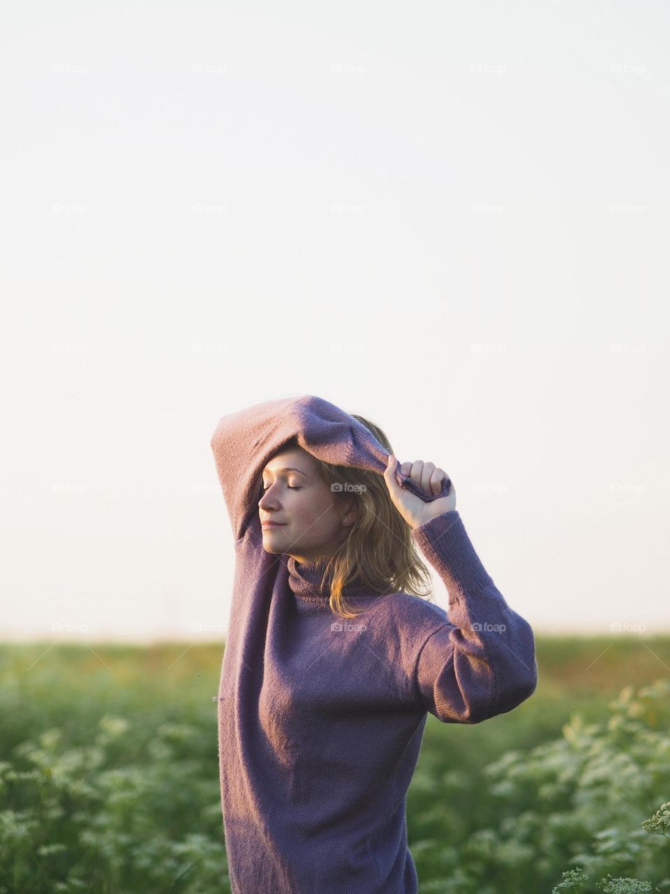 Young beautiful woman with blonde hair wearing lilac sweater standing on a green field in summer day, portrait of woman 