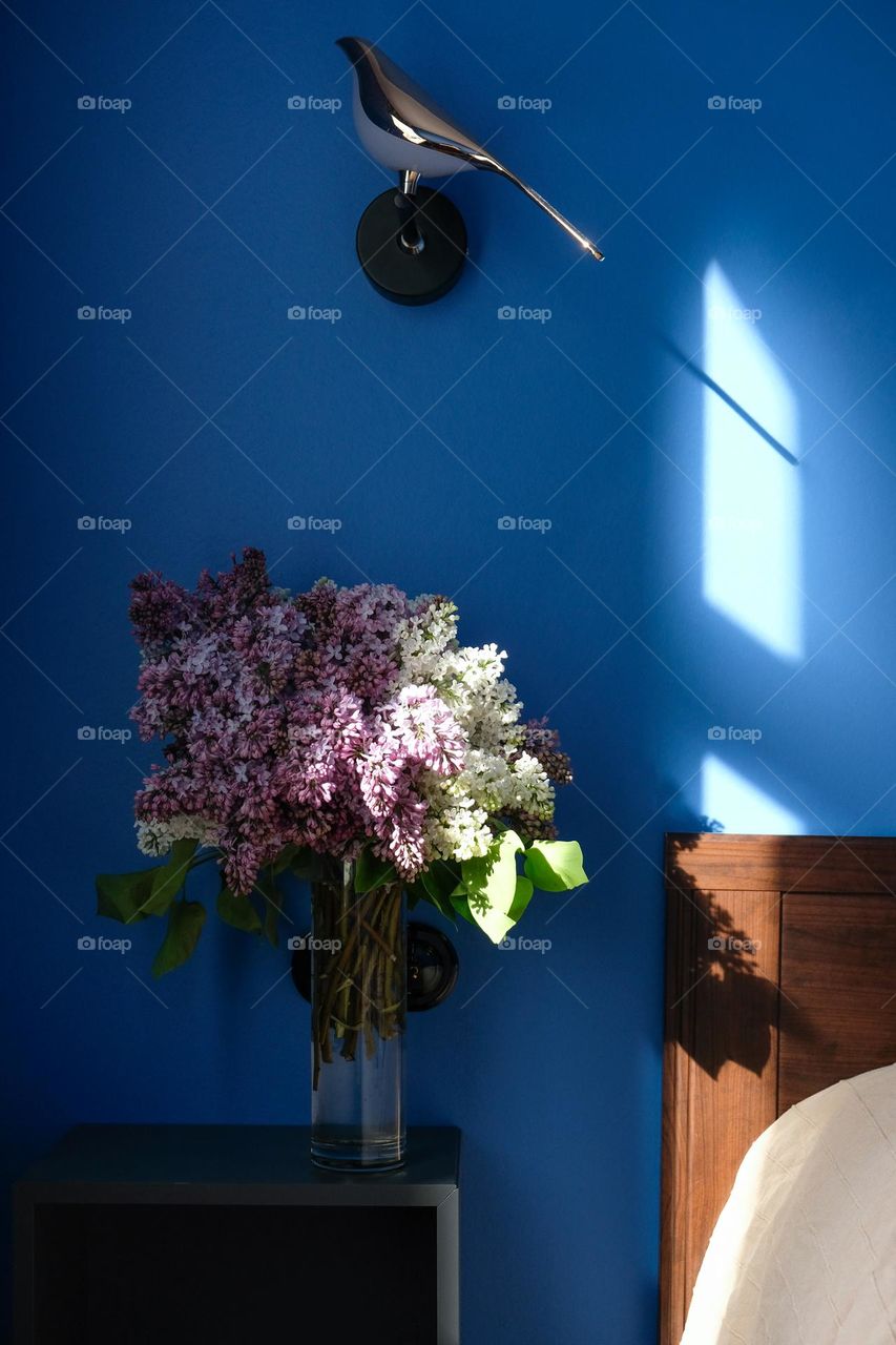 A bouquet of lilacs in the sunlight on a bedside table in a blue bedroom