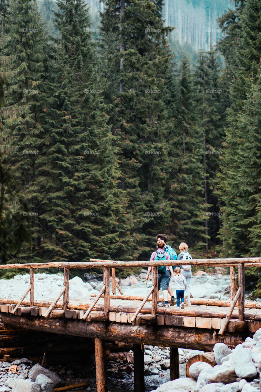 Family walking across a wooden bridge over a mountain stream. Spending vacation on wandering with backpacks in a mountains and forests