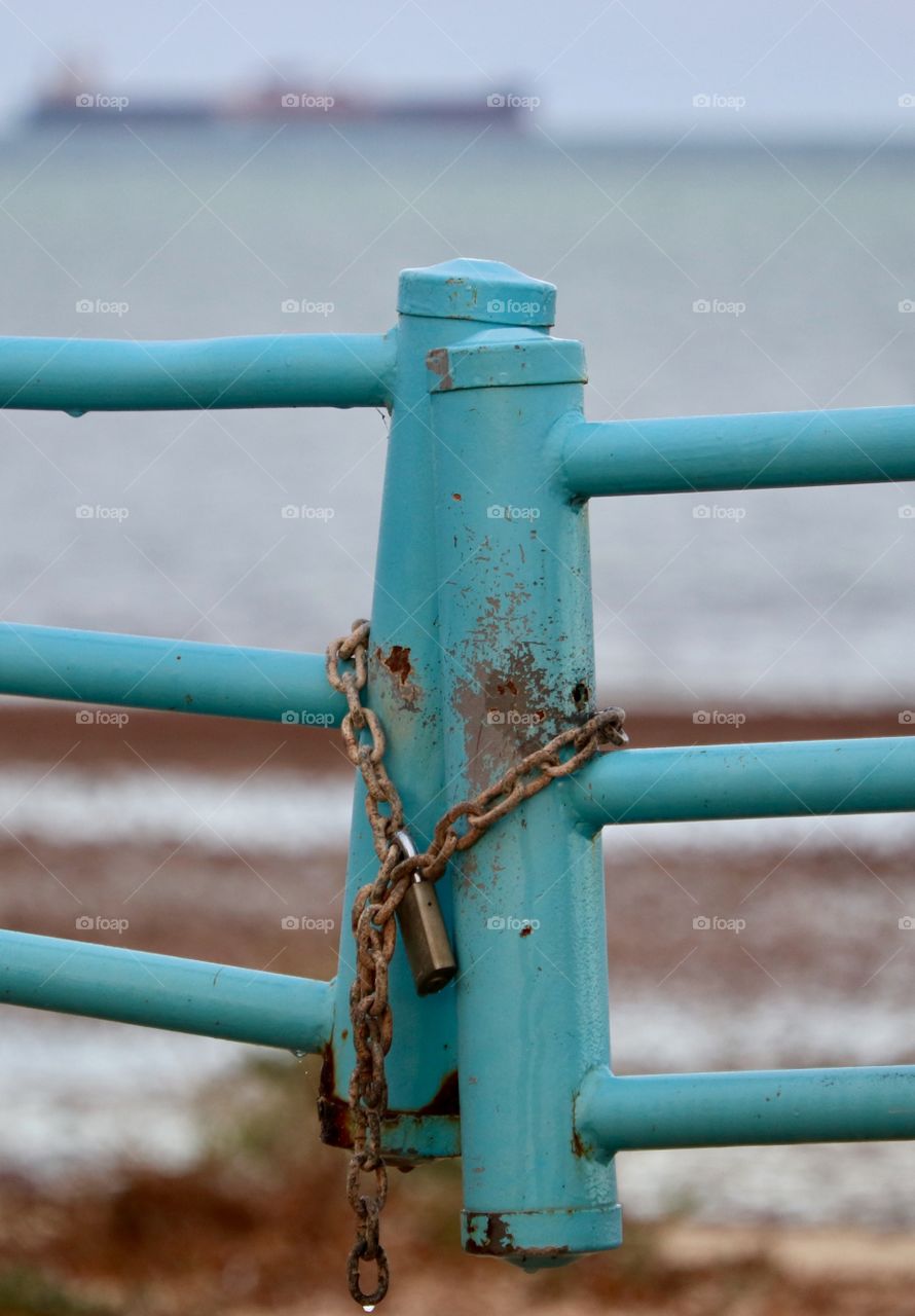 Closeup rusted padlock and chain attached to turquoise painted metal gate foreground with cargo ship on ocean horizon 