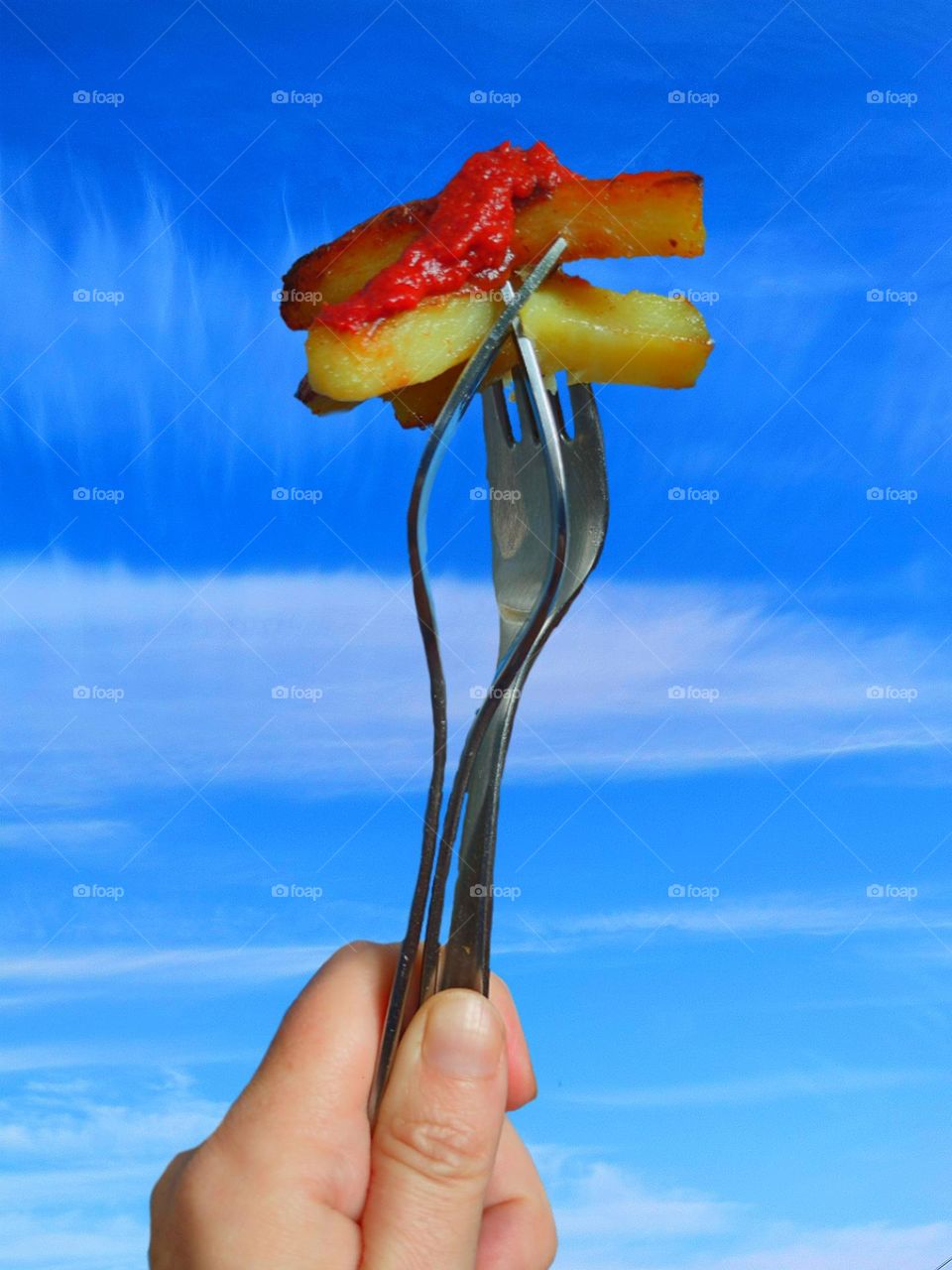 The hand holds three forks, on which are pieces of french fries and red ketchup on top. Blue sky background with white clouds