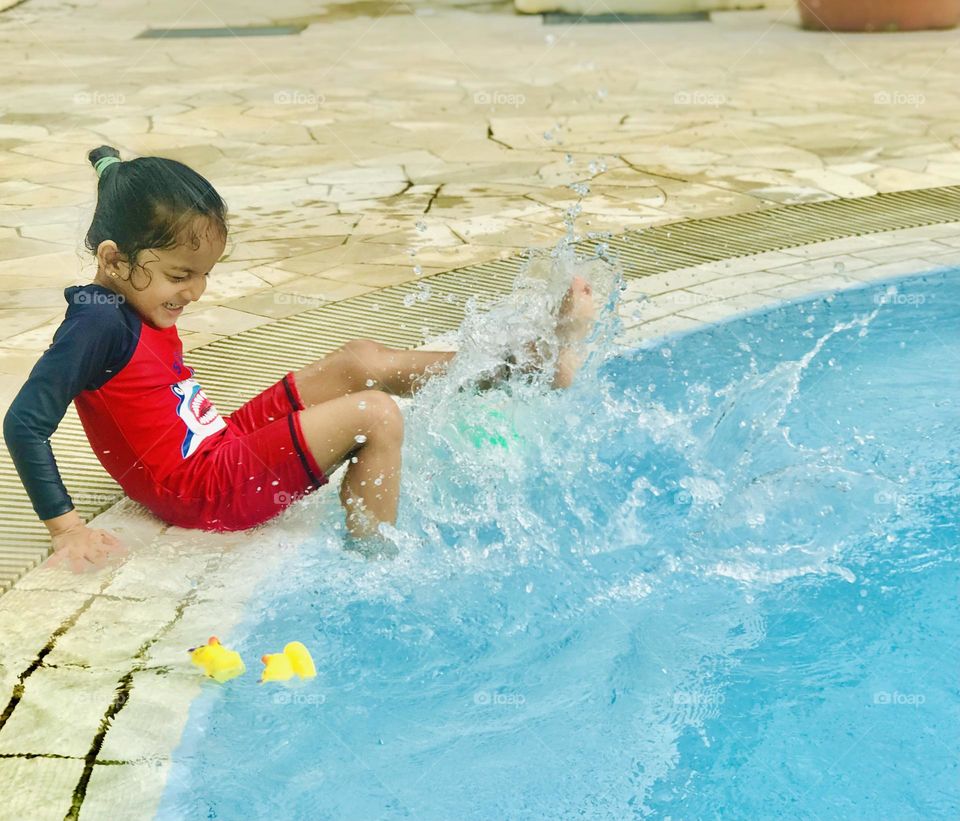 Everyone likes to go swimming pool in summer time,and 4 years old girl enjoying a lot at swimming pool by putting her legs in pool and moving to sprinkles water.