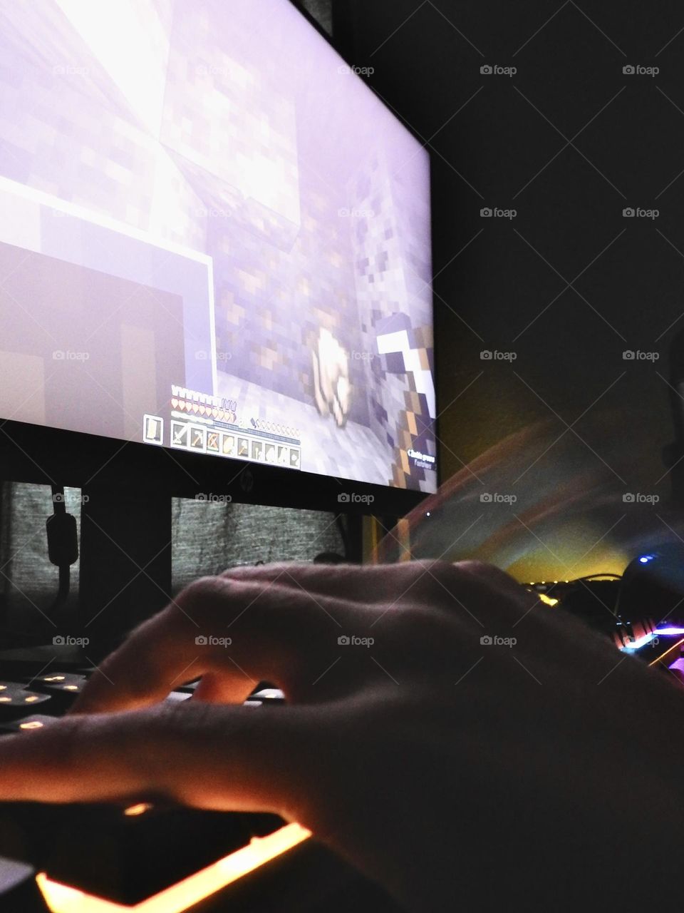 Teenager boy playing Minecraft on the gaming computer seen from his left side and close-up of his hand playing in a dark room.