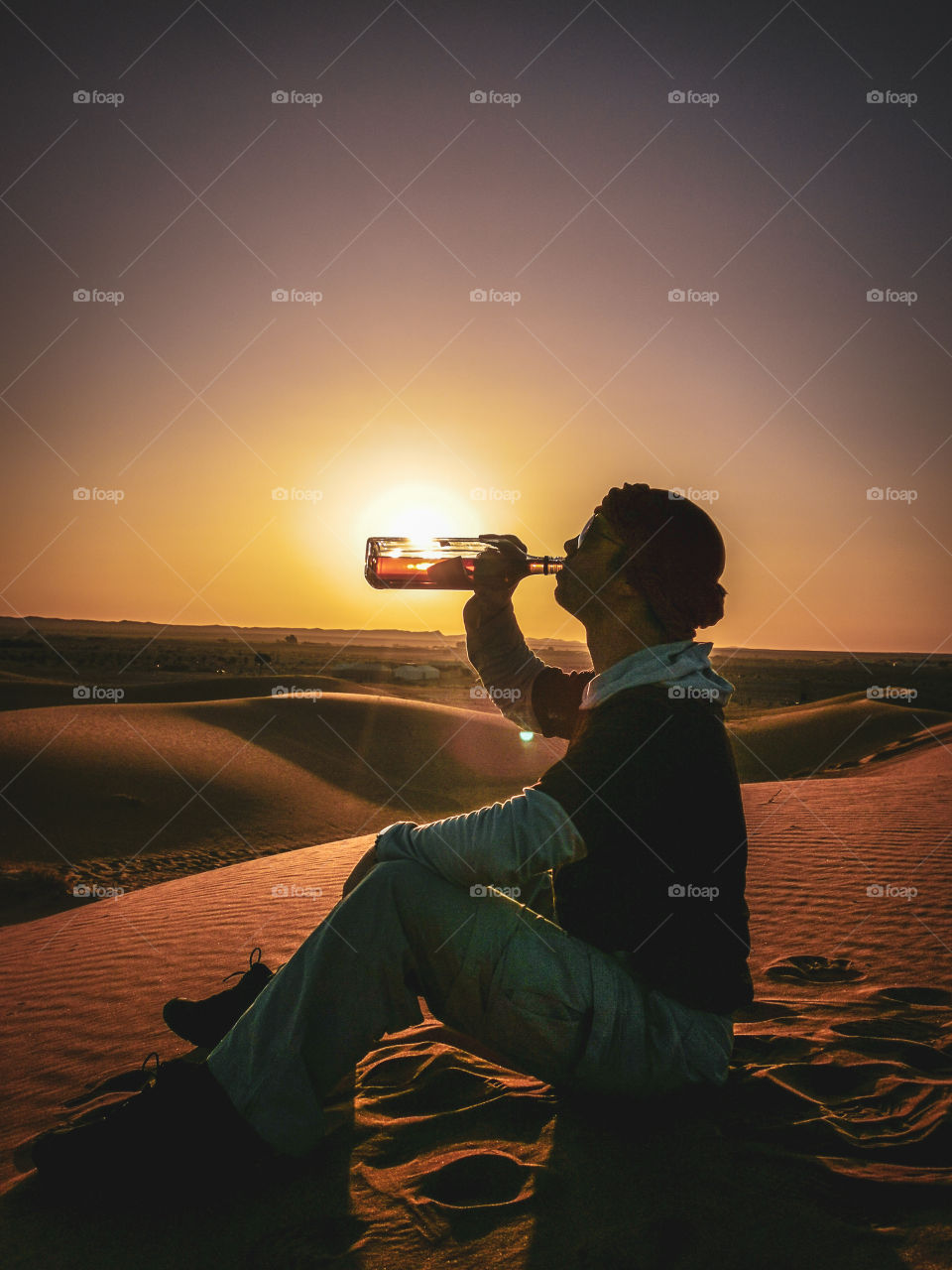 Young caucasian fit guy, drinking a whisky directly from the bottle during the sunsrise on the desert on Morocco.