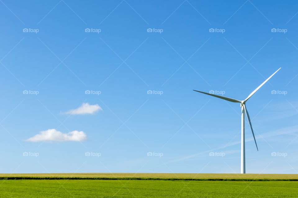 A wind turbine in yellow and green fields with two clouds on a blue sky