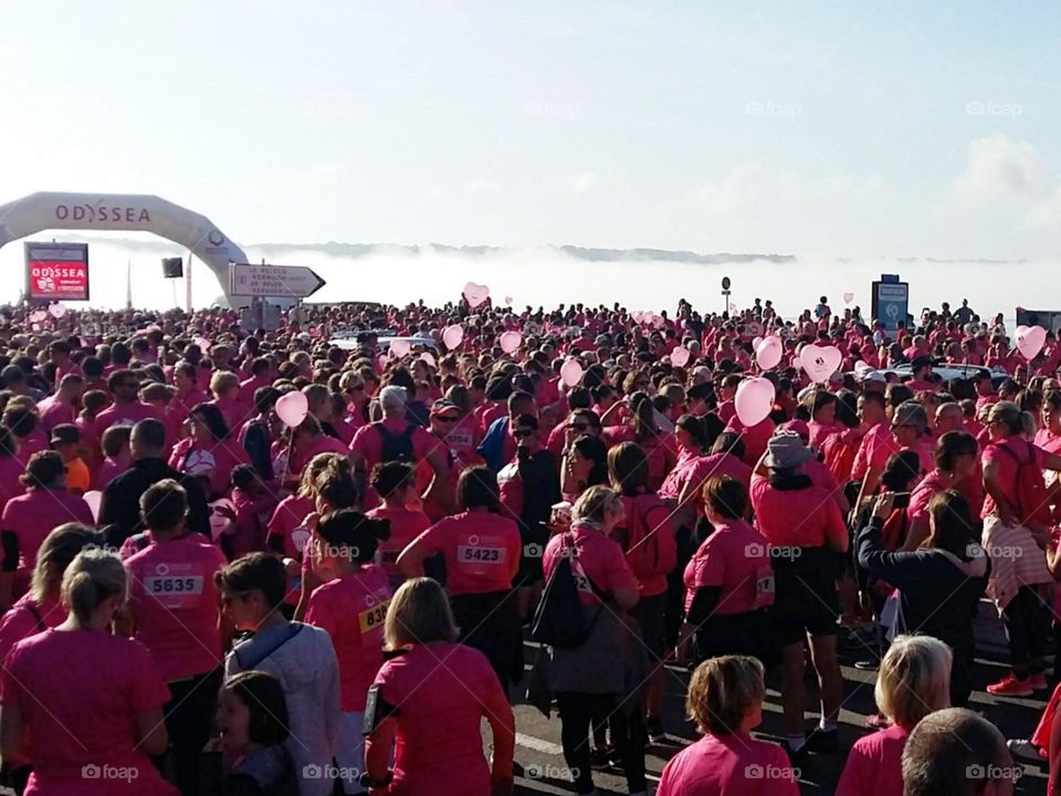 A crowd of athletes before the start of a race for breast cancer research. Everyone is wearing a pink t-shirt, some are racing with heart-shaped balloons.