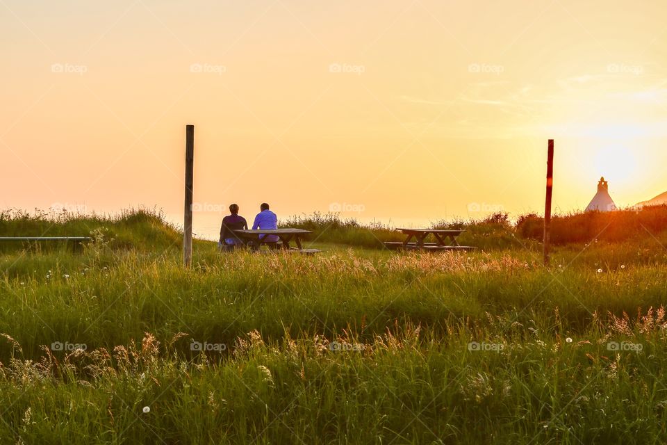 A couple of people in front of the camera sitting on a bench by the beach in the sunset 