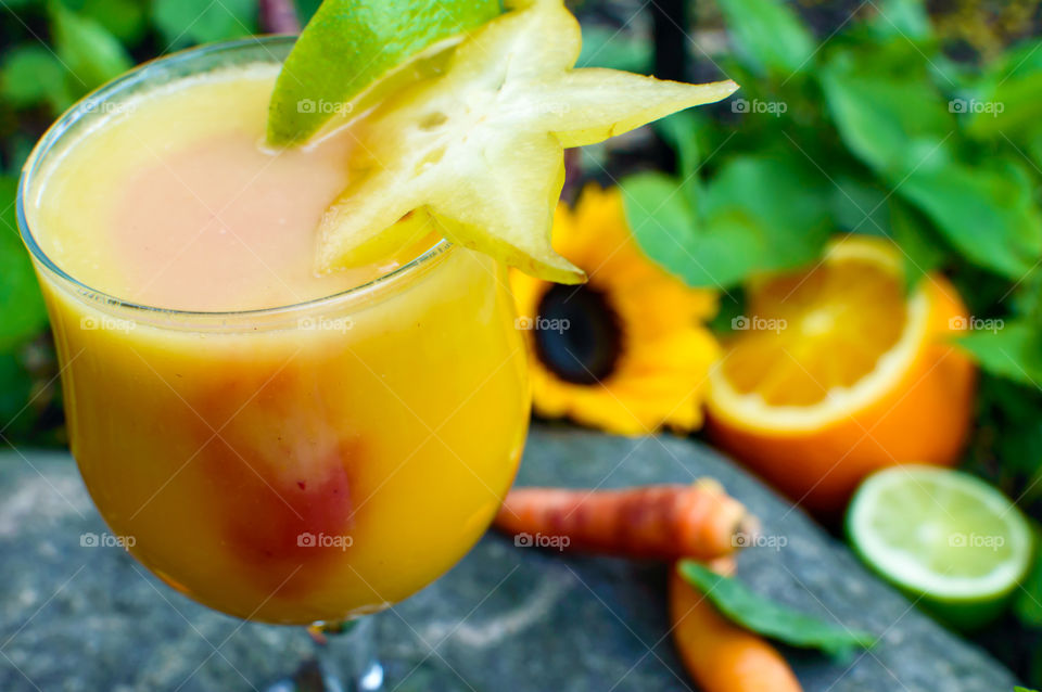 Orange lime mango and coconut fruit smoothie with red berry and carrot smoothie infusion in glass with ingredients in and sunflower in background decorated with slice of lime and star fruit