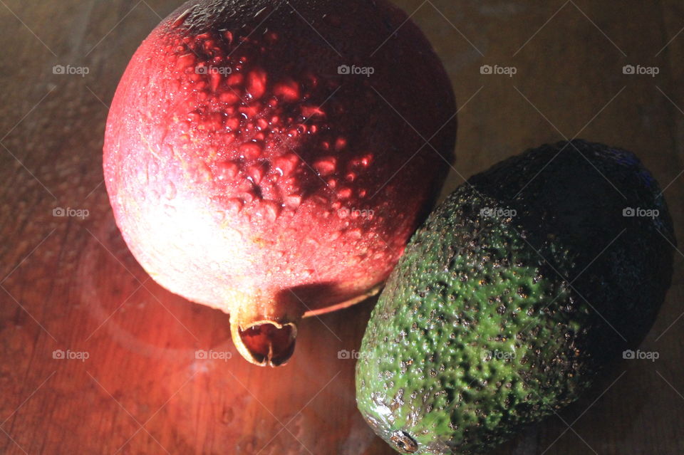 A red pomegranate and a green avocado still-life on a wood table. The fruit are wet and side-lit to show the droplets and textures of the fruit. 