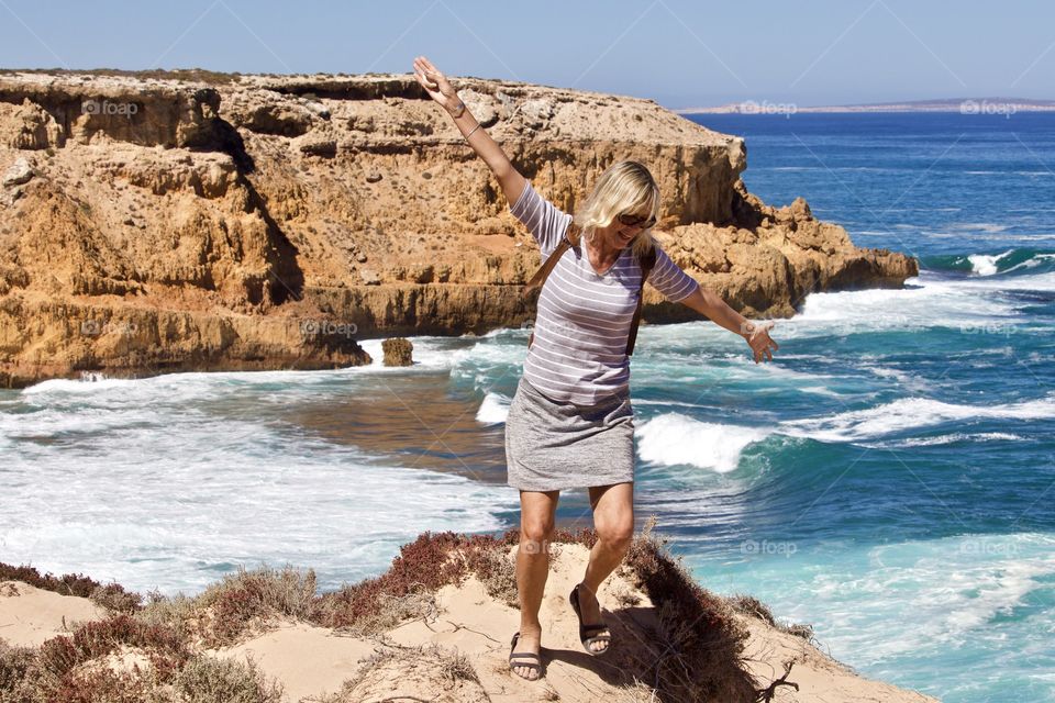 Playful blonde haired woman in skirt, top and backpack with outstretched arms on cliff top overlooking turquoise ocean and white water waves crashing to shore at Coffin Bay National Park remote beach in South Australia in summer 