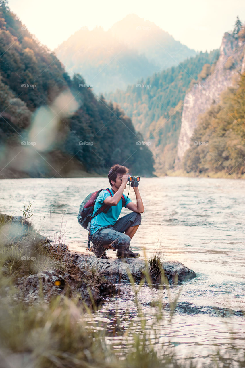 Young tourist with backpack looks through a binoculars on mountains peaks, stands on a rock over a river. Boy spends a vacation in mountains, wandering with backpack, he is wearing sports summer clothes