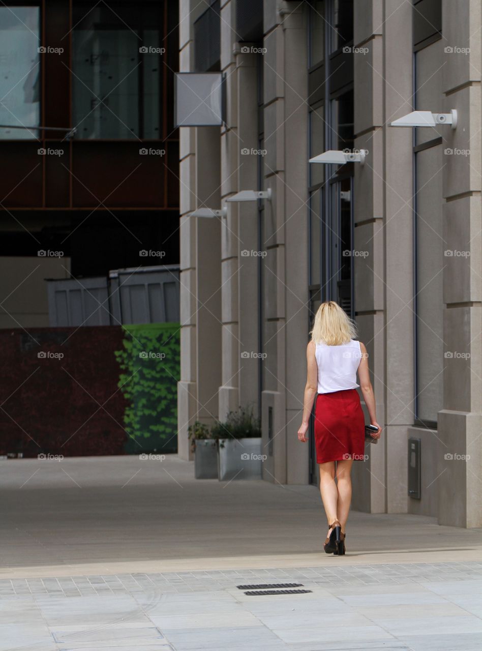 A blonde haired businesswoman walking along an alleyway in a red skirt and white blouse.