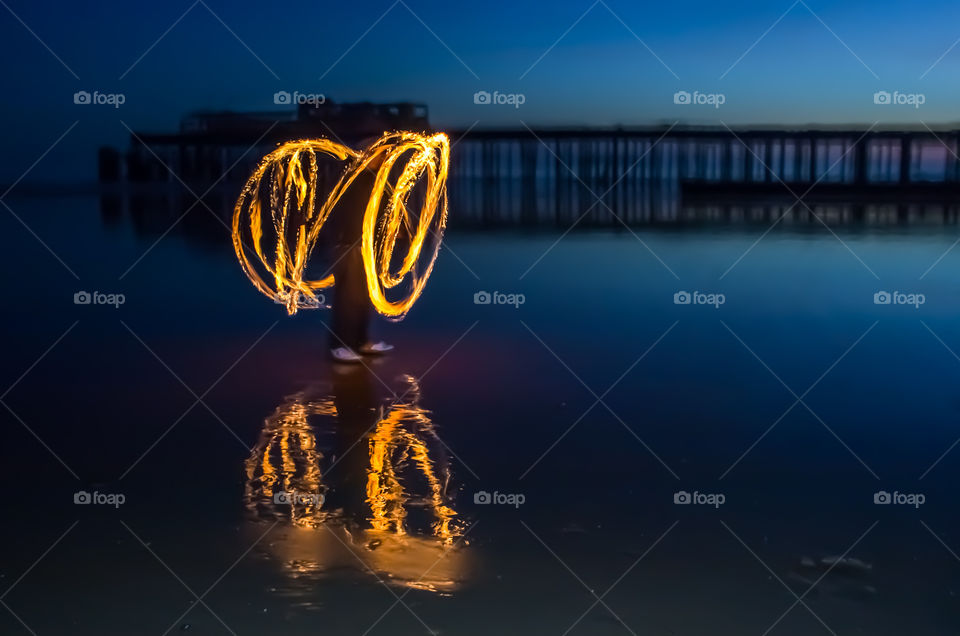 A person spinning fire poi, at dusk with a pier in the background, the scene is reflected on the wet beach