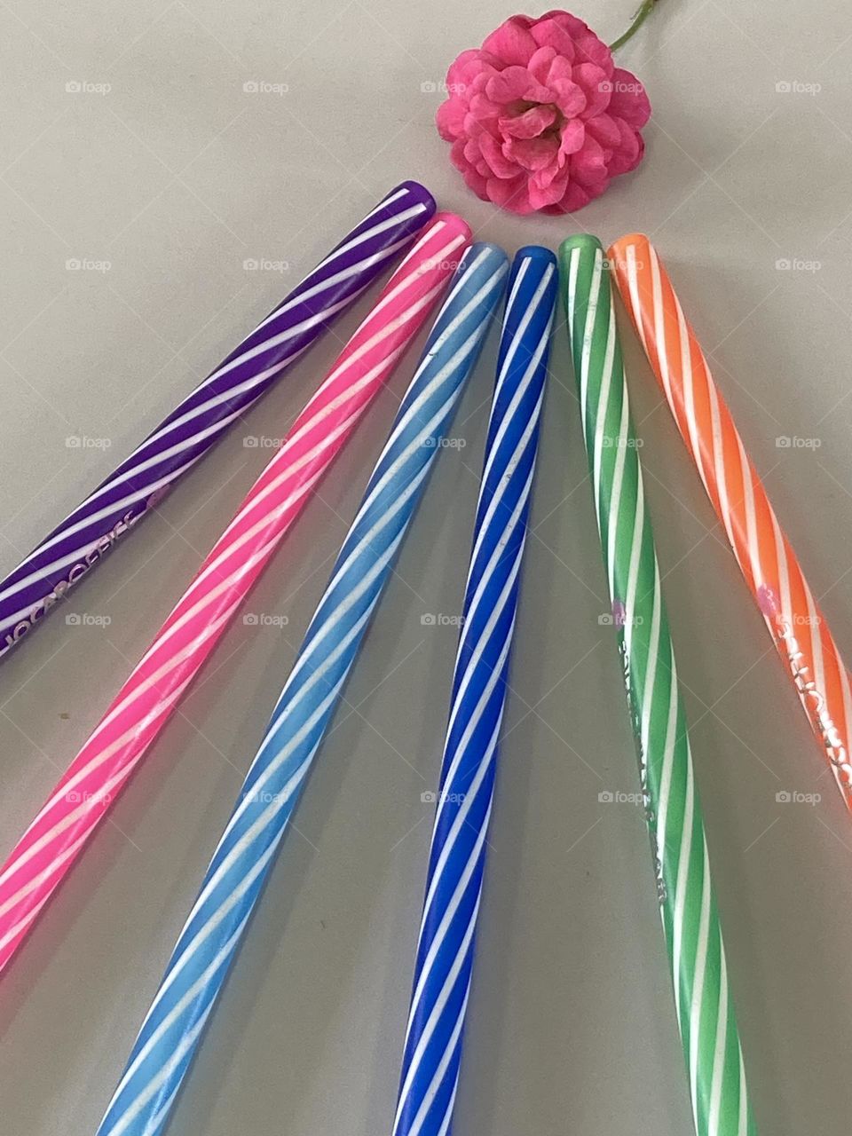 Composição com canetas coloridas, e para alegrar o cenário uma pequena rosa pink. Tudo sobre uma mesa cinza clara.