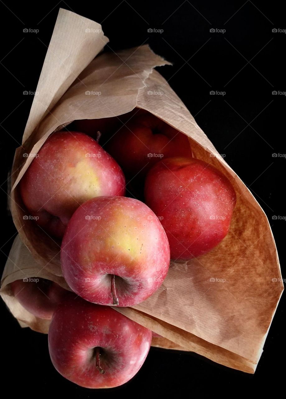 On a mirrored surface lies paper rolled up into a cone containing red apples. One apple has rolled out and is reflected in the mirror surface. Black background