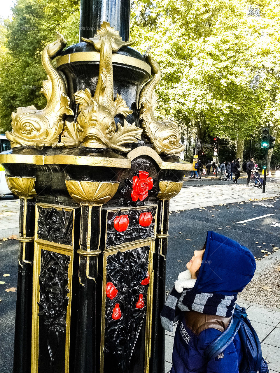 Una elegante farola en negro, rojo y dorado del paseo de Victoria de Londres  en la imagen brilla bajo una leve luz de sol invernal . El niño la mira atónito encandilado por su belleza y se olvida del viento que azota su cara tapada con capucha