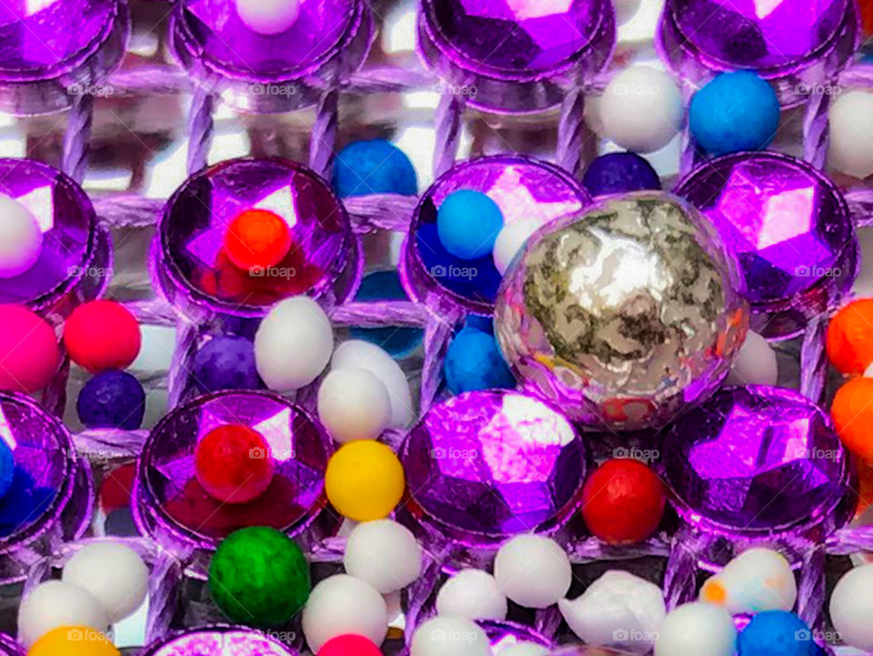 World in macro: Multi-coloured candy sprinkles & one silver candy dragee sitting on a purple gemstone net mesh over a mirror creates a multi-layered cornucopia of colour & reveals the textures & reflections of the silver dragees & purple gemstones. 