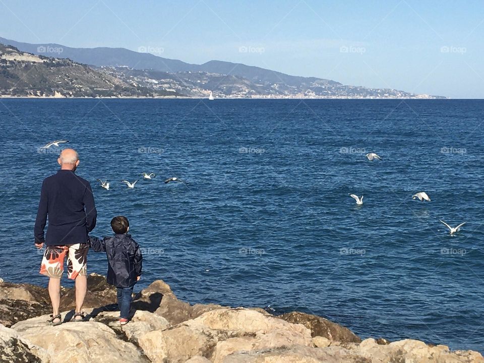 Old man with young boy observing blue sea and seagulls 