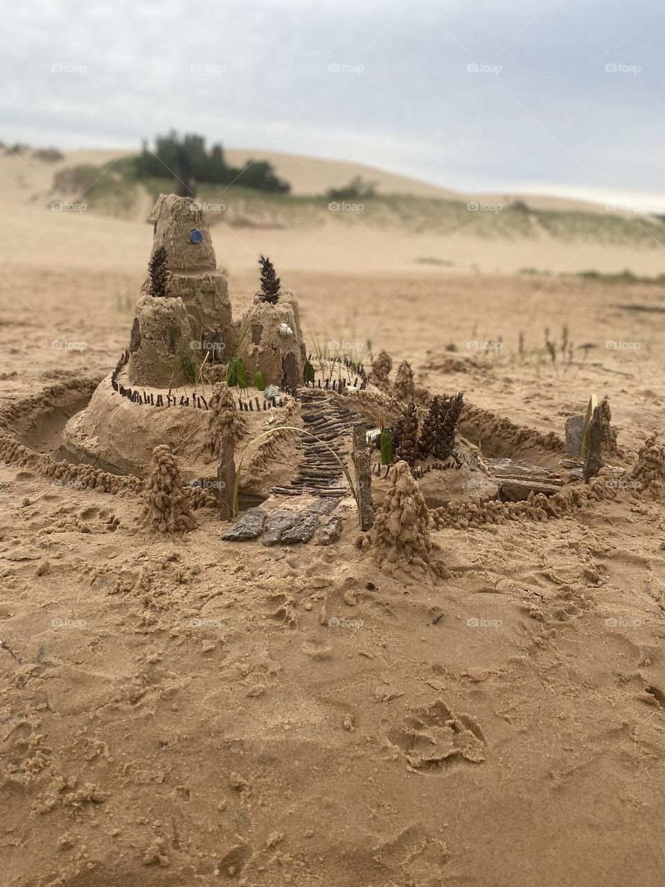 This image shows an intricate sandcastle on a beach, featuring multiple tiers and decorated with natural elements like pinecones, sticks, leaves, and shells. The sandcastle has a central tower surrounded by smaller structures, with detailed pathways 