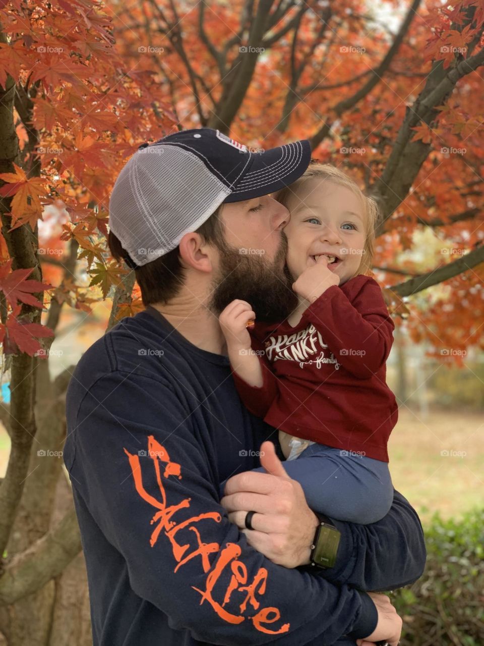 A dad kissing his baby girl in the fall in front of a beautiful colorful tree. 