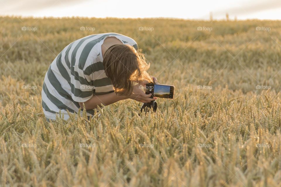 a teenager photographs a sunset in a wheat field on a phone camera that is mounted on a tripod