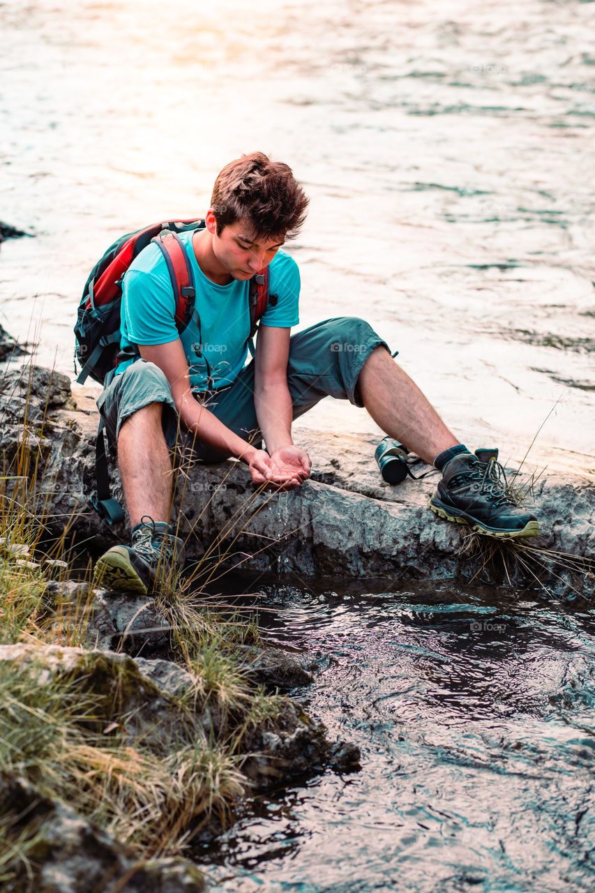 Young boy takes pure water from a river and holds it in the hands. He is sitting on a rock over the river, rests during a hike, spends a vacation on wandering with backpack, he is wearing sport summer clothes