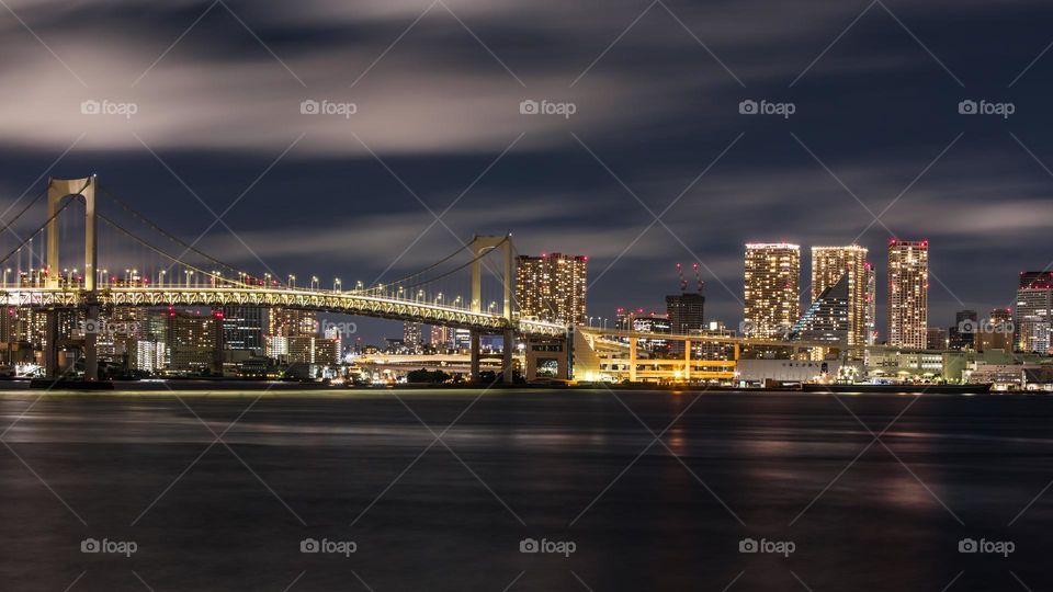 Rainbow Bridge over Tokyo Bay at night captured in long exposure. Movements of clouds and water. Office, business, and residential high rise buildings make the skyline of this outer parts of the Japanese capital city.