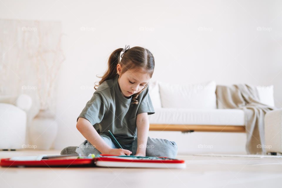 Preschool little girl is sitting on the floor and drawing at bright room at home