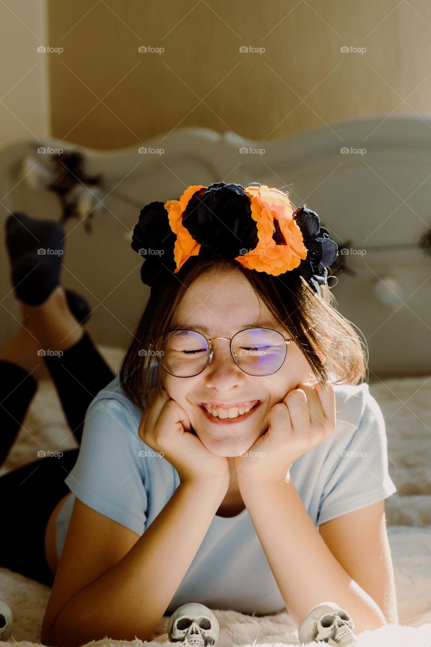 Portrait of one beautiful happy Caucasian girl with a Halloween headband, closing her eyes, cutely lying on the bed in the bedroom on an autumn sunny day, close-up side view.