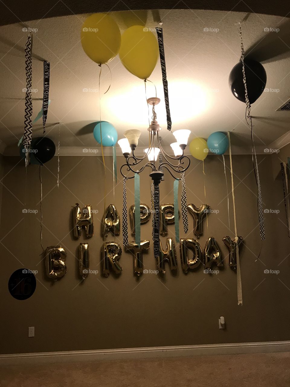 A shiny and golden Happy Birthday balloon banner hanging up on the wall surrounded by a bunch of yellow, blue and black helium party balloons and streamers. Decorating for a sweet sixteen birthday party. 