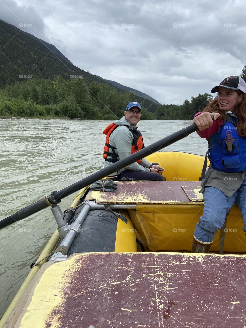 A strong woman rowing a raft during a tour in Alaska while a man is relaxing in the background.