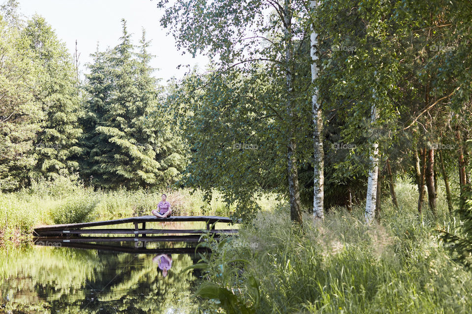 Woman sitting on a bridge over a lake, among the trees, close to nature, during summer vacations. Candid people, real moments, authentic situations