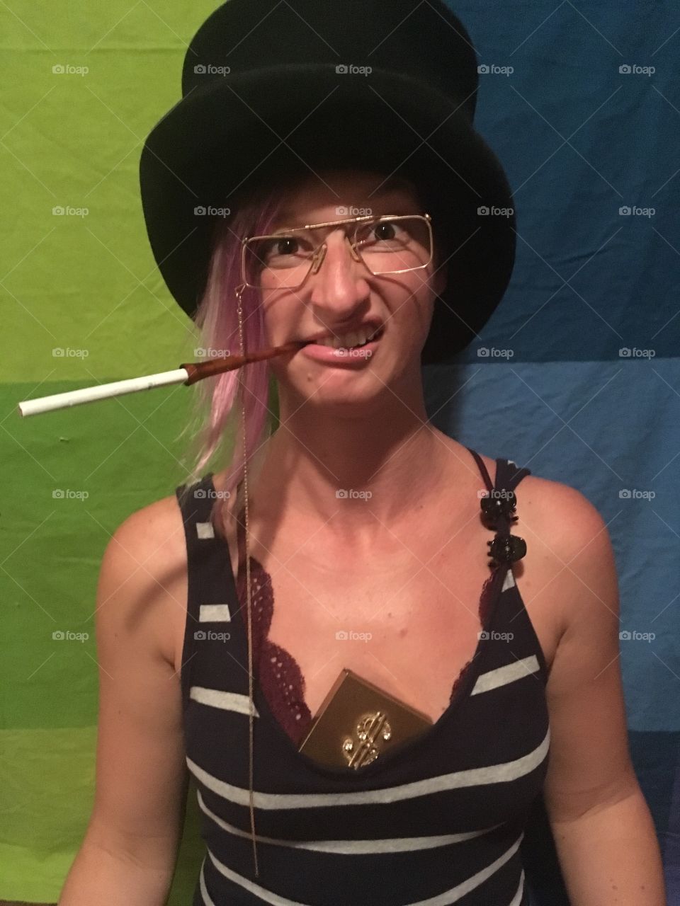 Woman making a silly snarling face with cigarette in a top hat and glasses in front of a colorful green and blue background