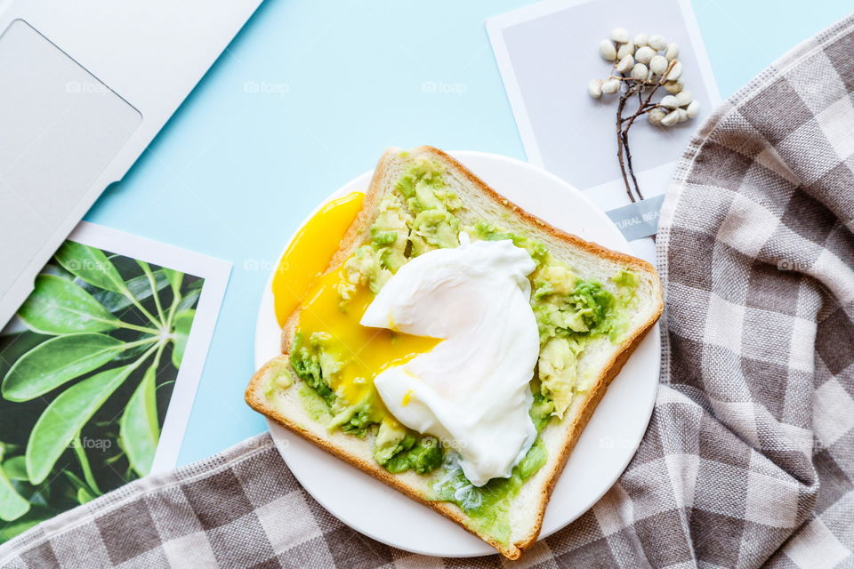 Sandwich with fresh green avocado, poached egg on white plate lying on blue background 