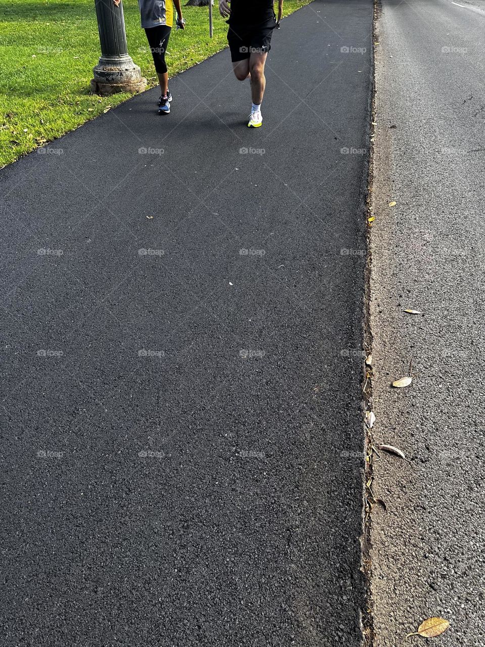 Lower half shown of a man and woman jogging down a freshly paved asphalt path near green field and a road