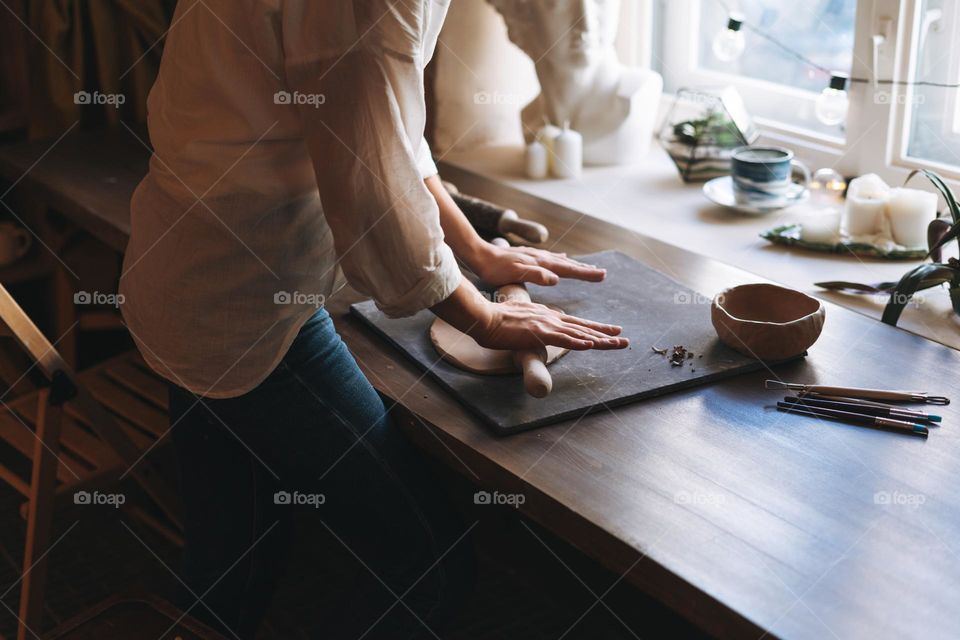 Young attractive woman in white shirt ceramic artist decorating clay plate with tool at table in pottery workshop. Handmade work student, freelance small business