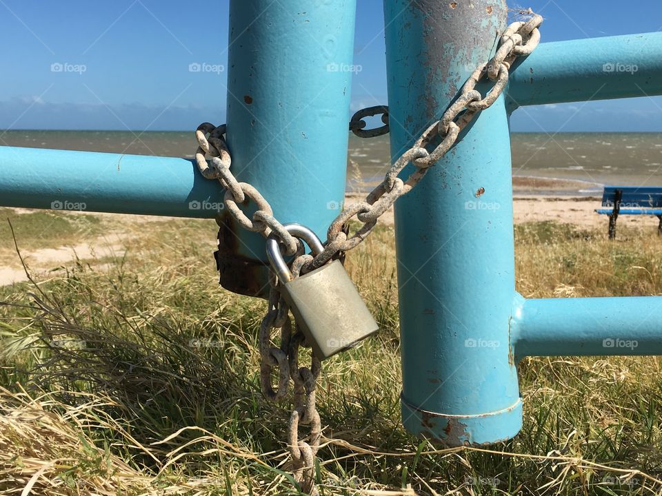A steel grey padlock and chain wrapped around blue pole of gate with ocean backdrop
