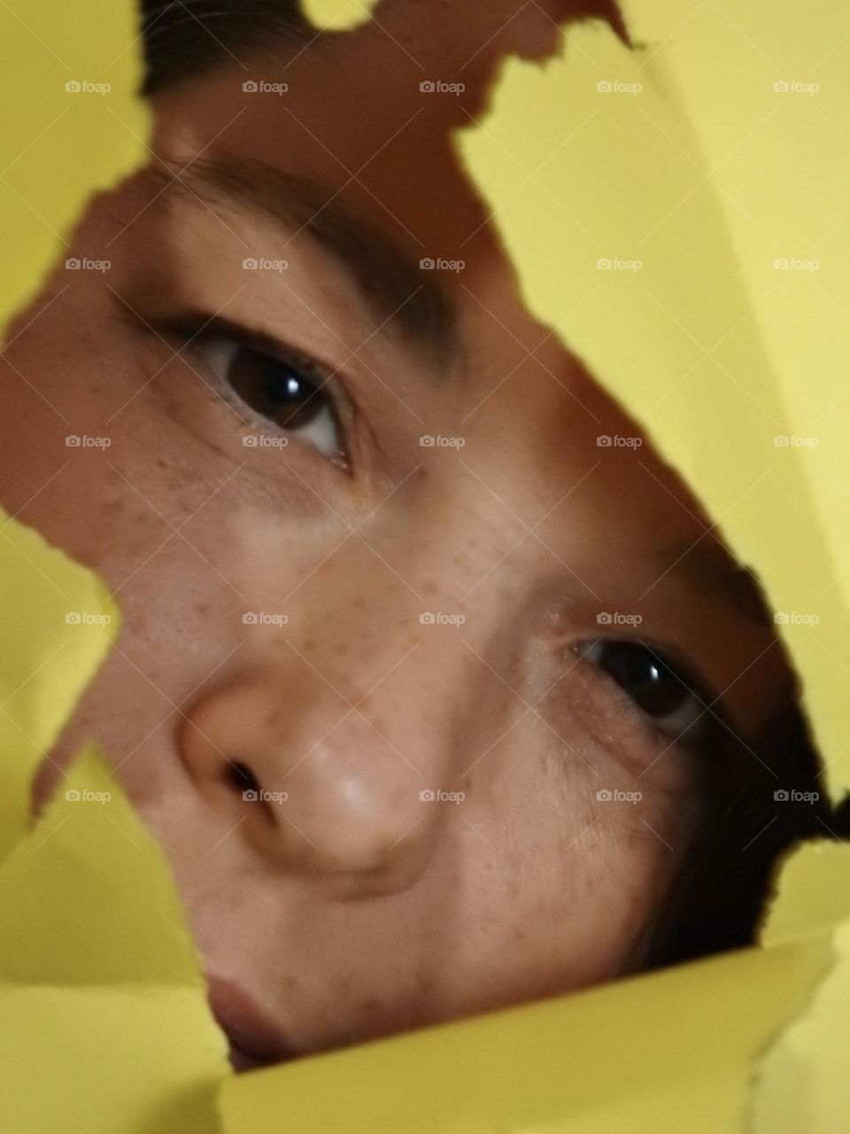 Face skin in the forties of a woman with freckles and brown hair seen through a broken yellow paper.