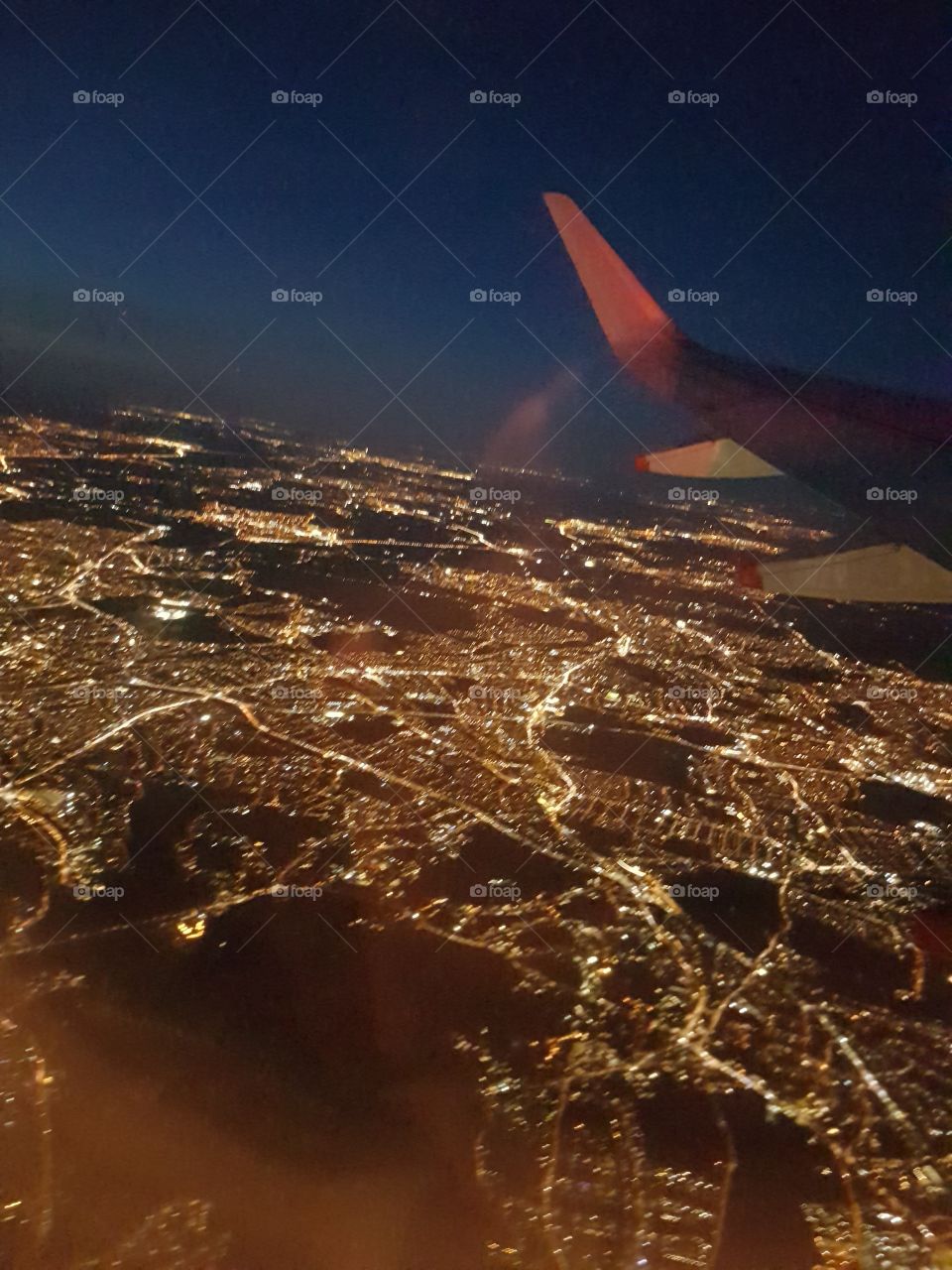 Evening flight, flying over glow of city lights on earth below, looking through jet plane window with wing in view
