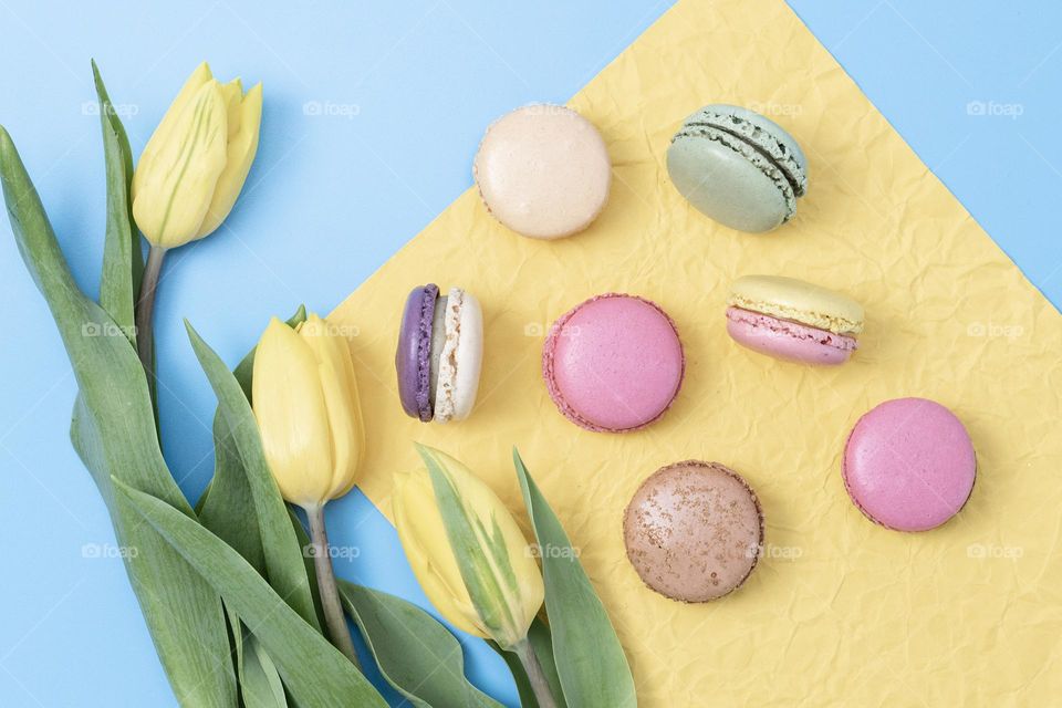 Appetizing pastel-colored macaroon dessert with a bouquet of yellow tulips lies on a crumpled yellow napkin on a light blue background, flat lay close-up.