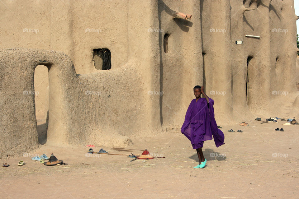 A girl wearing a purple robe walks past a mosque near Djenne, Mali