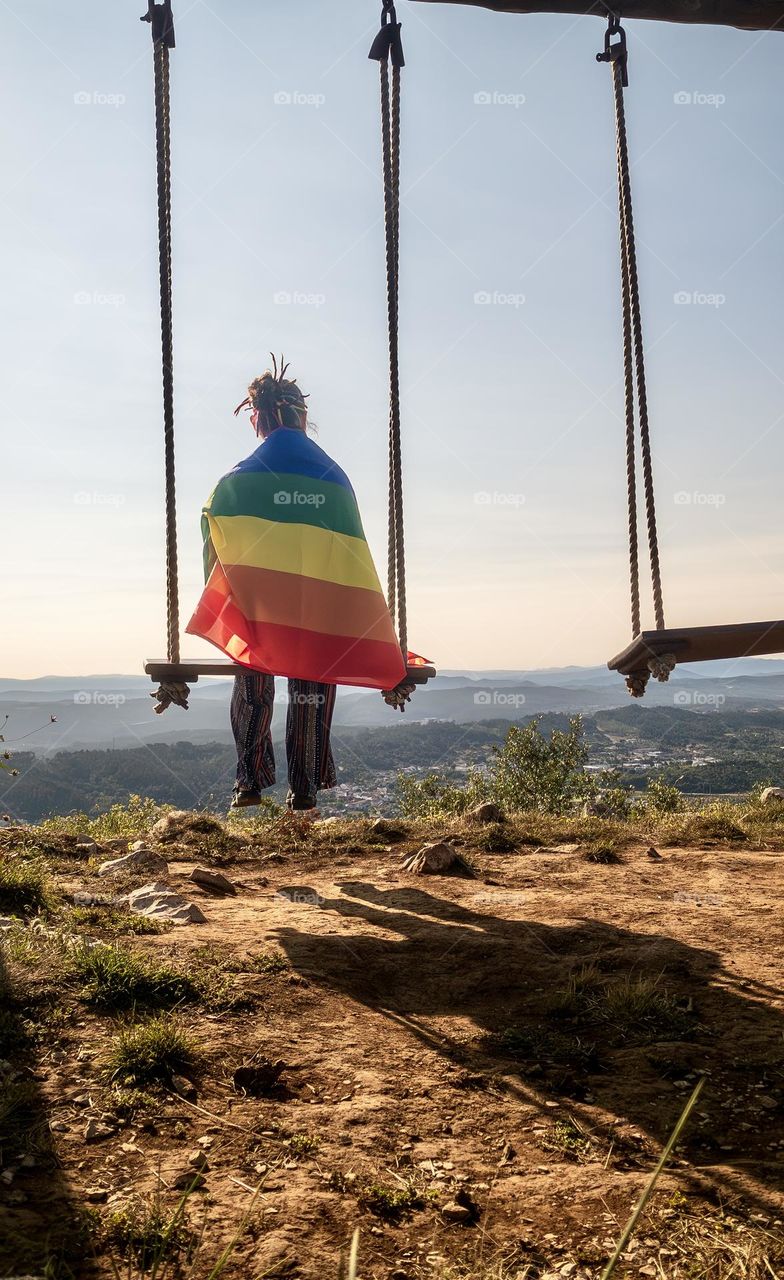 A person sitting on a large swing, overlooking the landscape, draped in a rainbow coloured flag