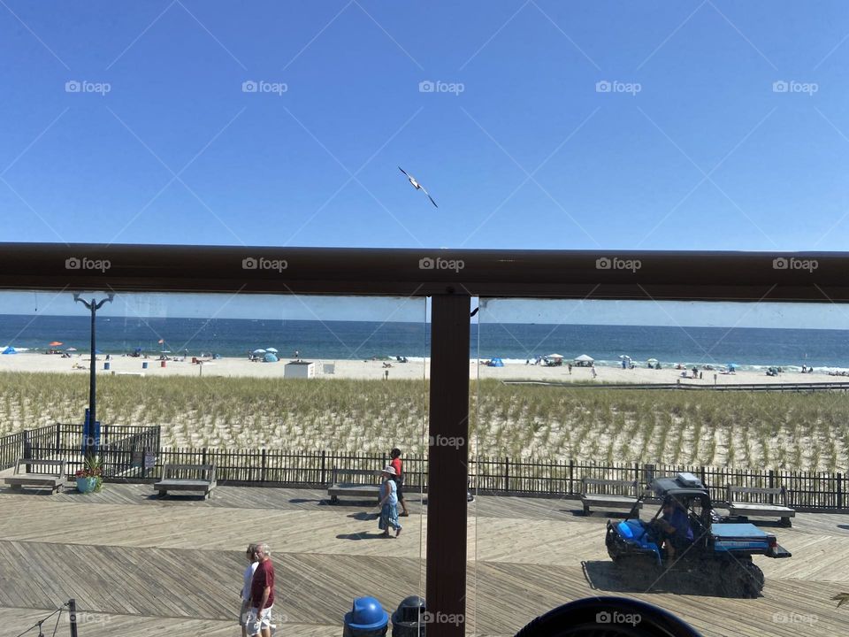 People walking on the boardwalk in Seaside Heights, NJ on a September day. The sky is blue, the ocean even bluer, and a Seagull soars above the railing. I took this from an upstairs Mexican restaurant on the boardwalk called Spicy Cantina.
