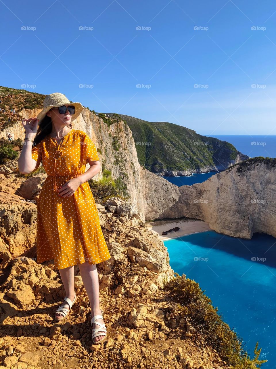 A young beautiful Caucasian girl in a yellow polka-dot dress, straw hat and sunglasses standing on the edge of a cliff overlooking the sea, a deserted beach with a beached pirate ship on a clear sunny summer day, close-up view from the side.