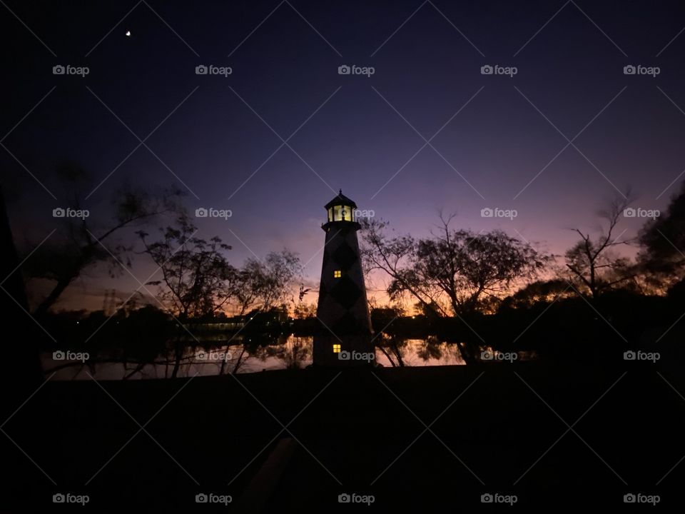 A Beautiful Sun-filled Day Clear Sky’s Overhead this Lighthouse poised for Bright Moonlit Night Filled By Night     Colors. Reflections from Peaceful Lake Water with Backlit Twilight hanging on into Night. 