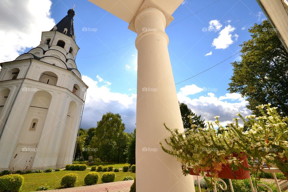 view from the porch of the monastery building. a huge white column of lawn flowers and an old bell tower. blue sky and bright sun.