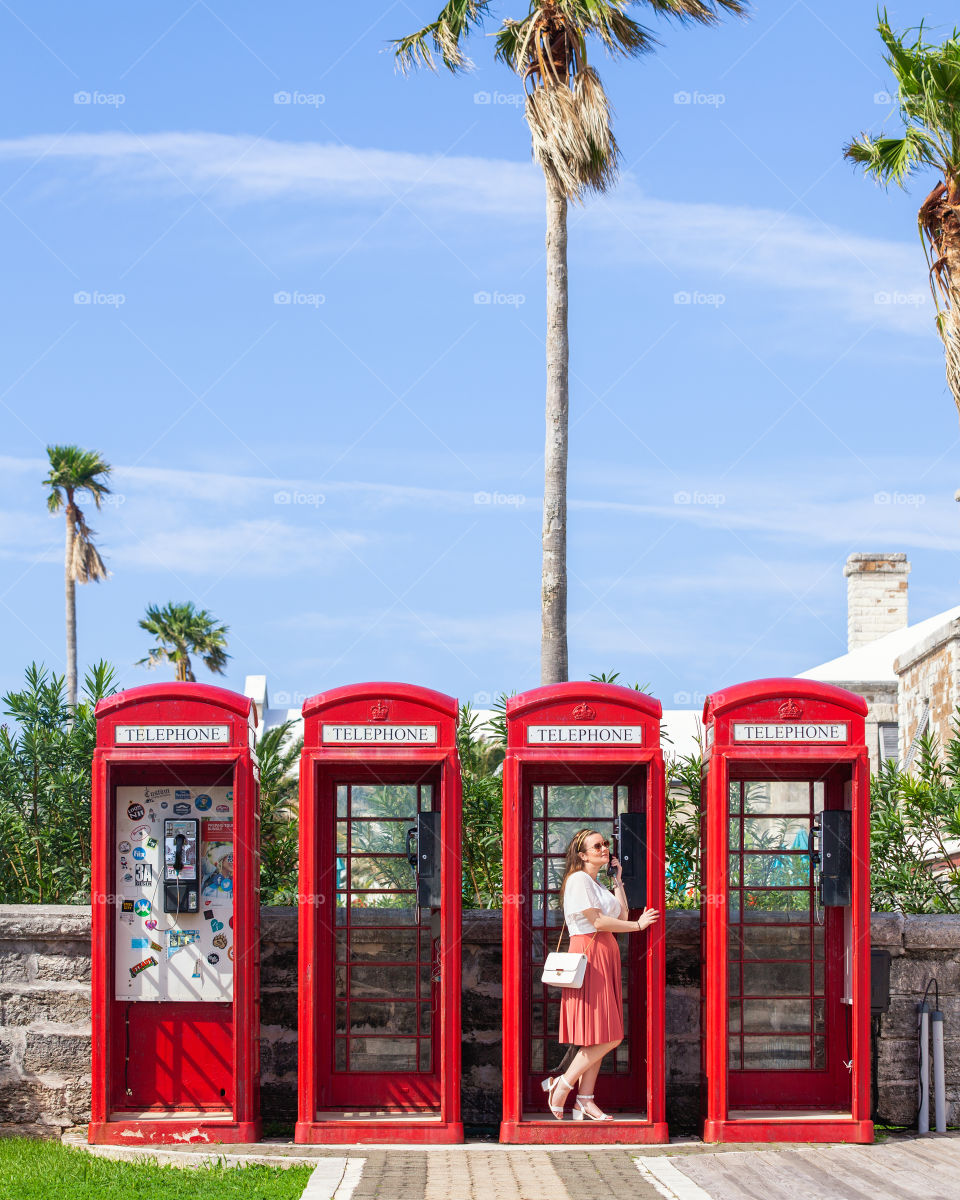 Young attractive female in light pink and white summery outfit standing in vibrant red telephone boxes, surrounded by lush greenery and palm trees under a blue sky.