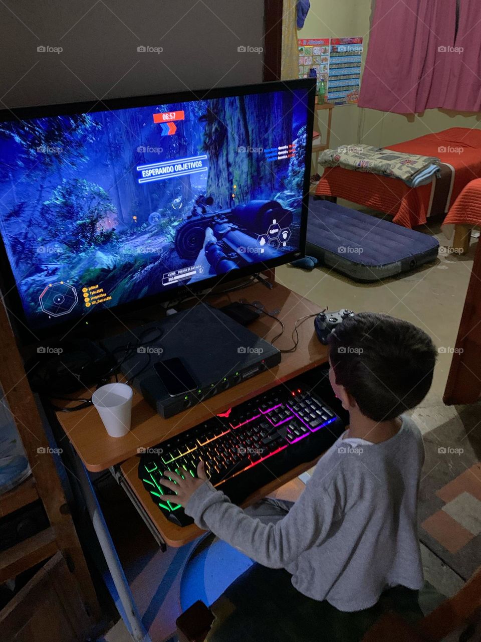 Little boy sitting on a desk, playing a shooter game in a computer with keyboard and mouse.