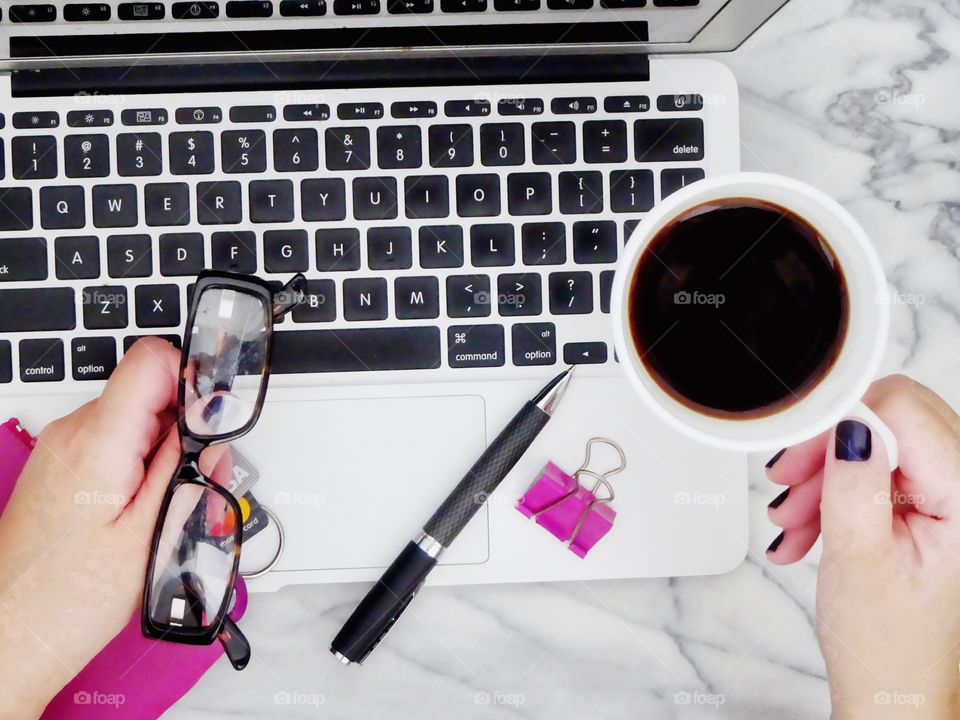 Flat lay overhead shot of laptop, woman's hands holding a cup of coffee and credit cards in pink wallet, glasses, pen and pink paper clip, on a marble table - ready to do some online shopping. 